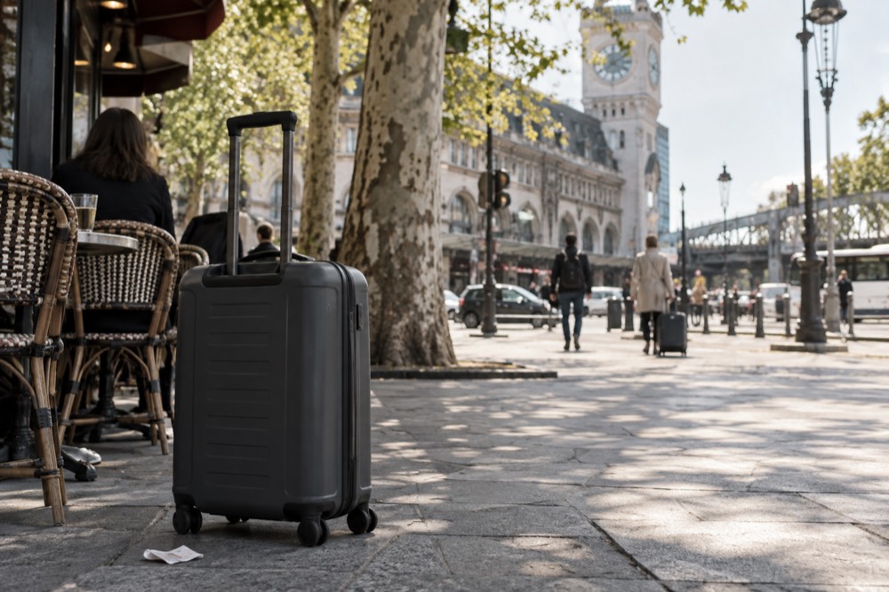 A suitcase on a wide sunlit Paris pavement outside a corner cafe terrace