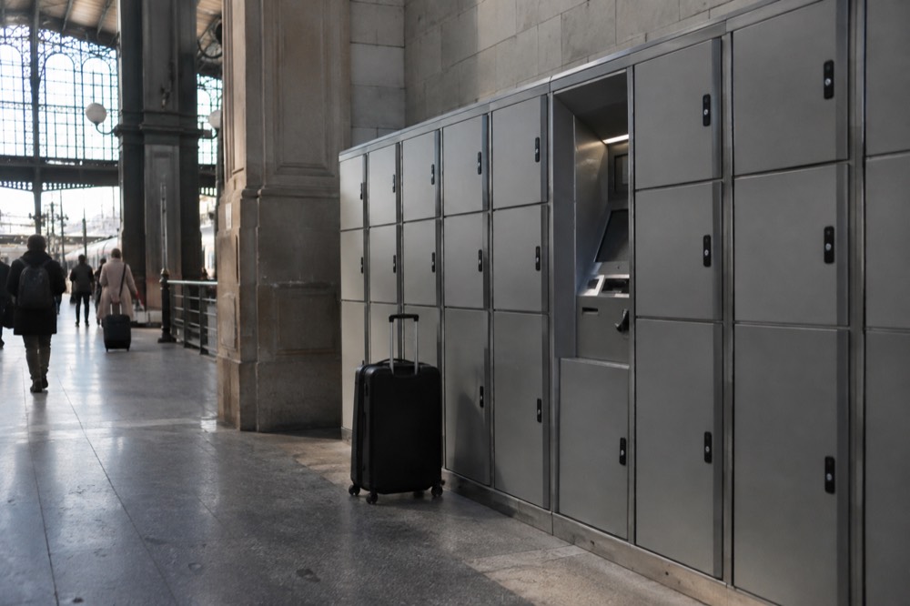 Automated luggage lockers in a row along a station wall with a trolley case parked beside them