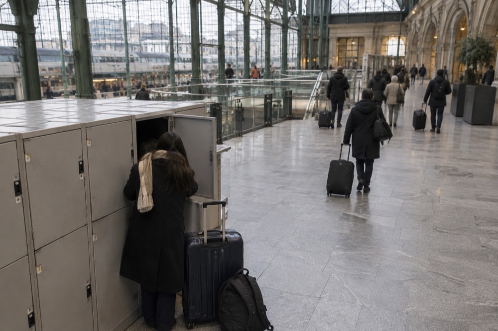 Railway station concourse with a left-luggage locker area and travellers passing with trolleys