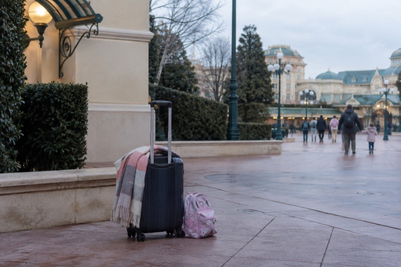 A wheeled suitcase and a child's backpack set down on the wide paved esplanade at Marne-la-Vallee east of Paris, where families reach Disneyland Paris before their rooms are ready.