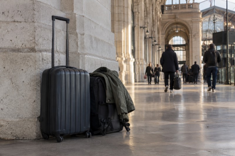 A suitcase and a smaller bag set down against a stone column on the concourse of a Paris mainline station, near the street exits where the shops and hotels that take bags are found.