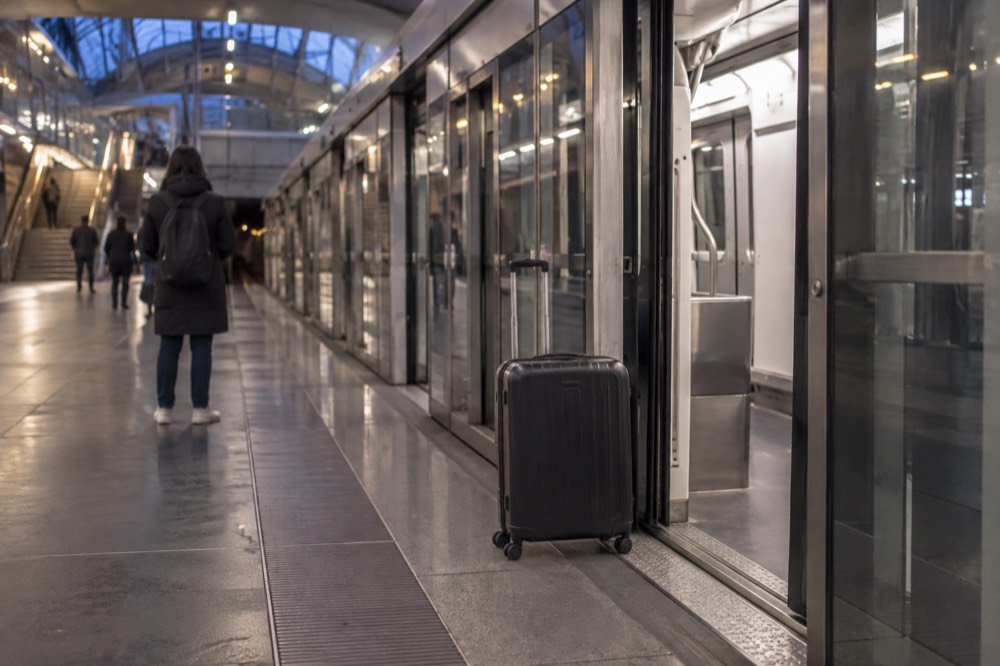 A modern metro platform with a lift door open and a wheeled suitcase beside a traveller waiting for a driverless train