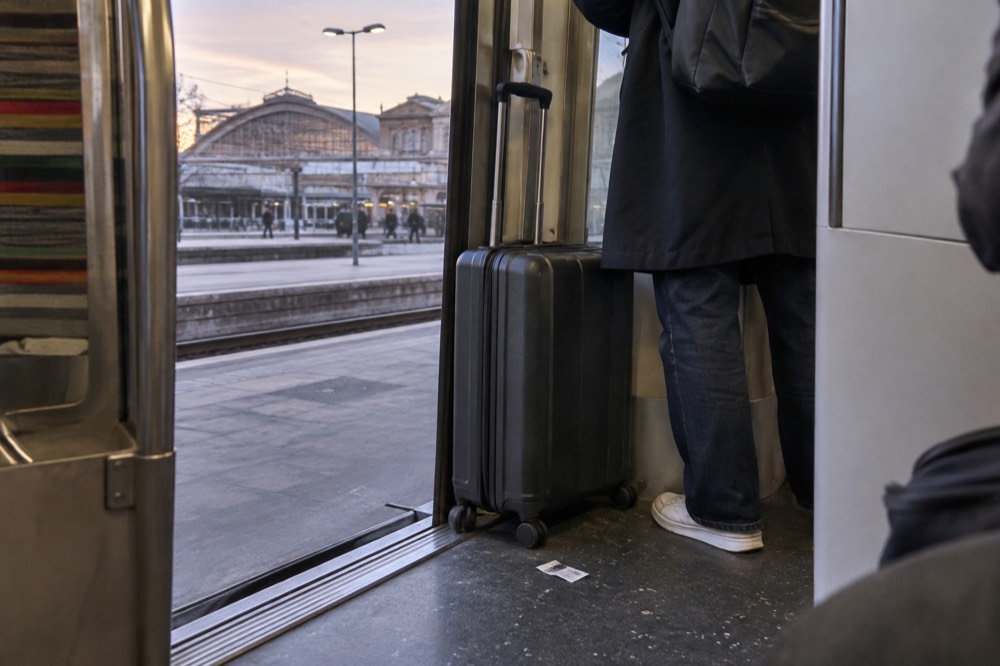 The interior of a commuter train with a wheeled suitcase held steady in the doorway well beside a standing passenger