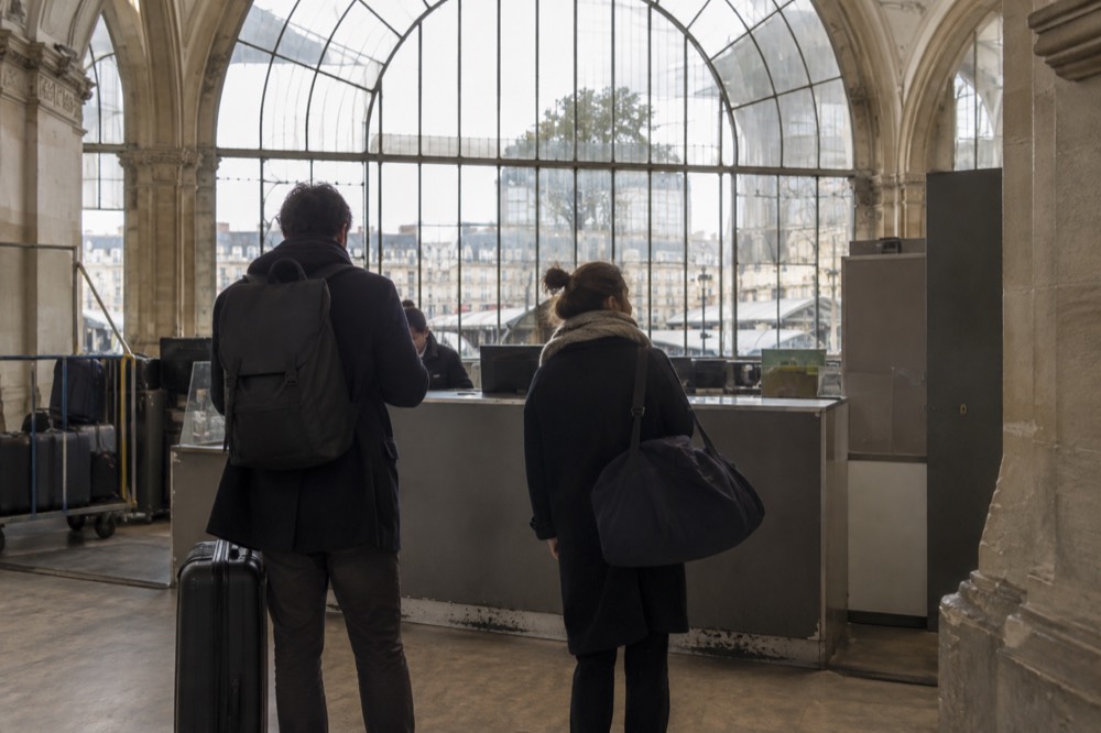 A staffed left luggage counter in a Paris station with a queue of two travellers.